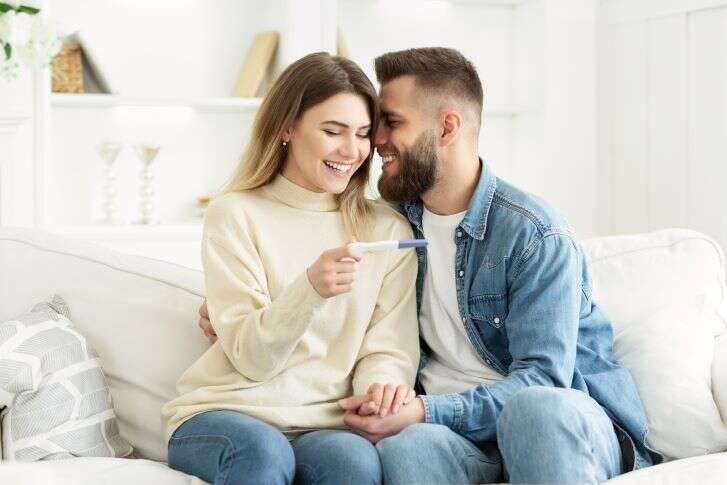 Eine Frau und ein Mann sitzen auf einer Couch und blicken erfreut auf einen Schwangerschaftstest.
